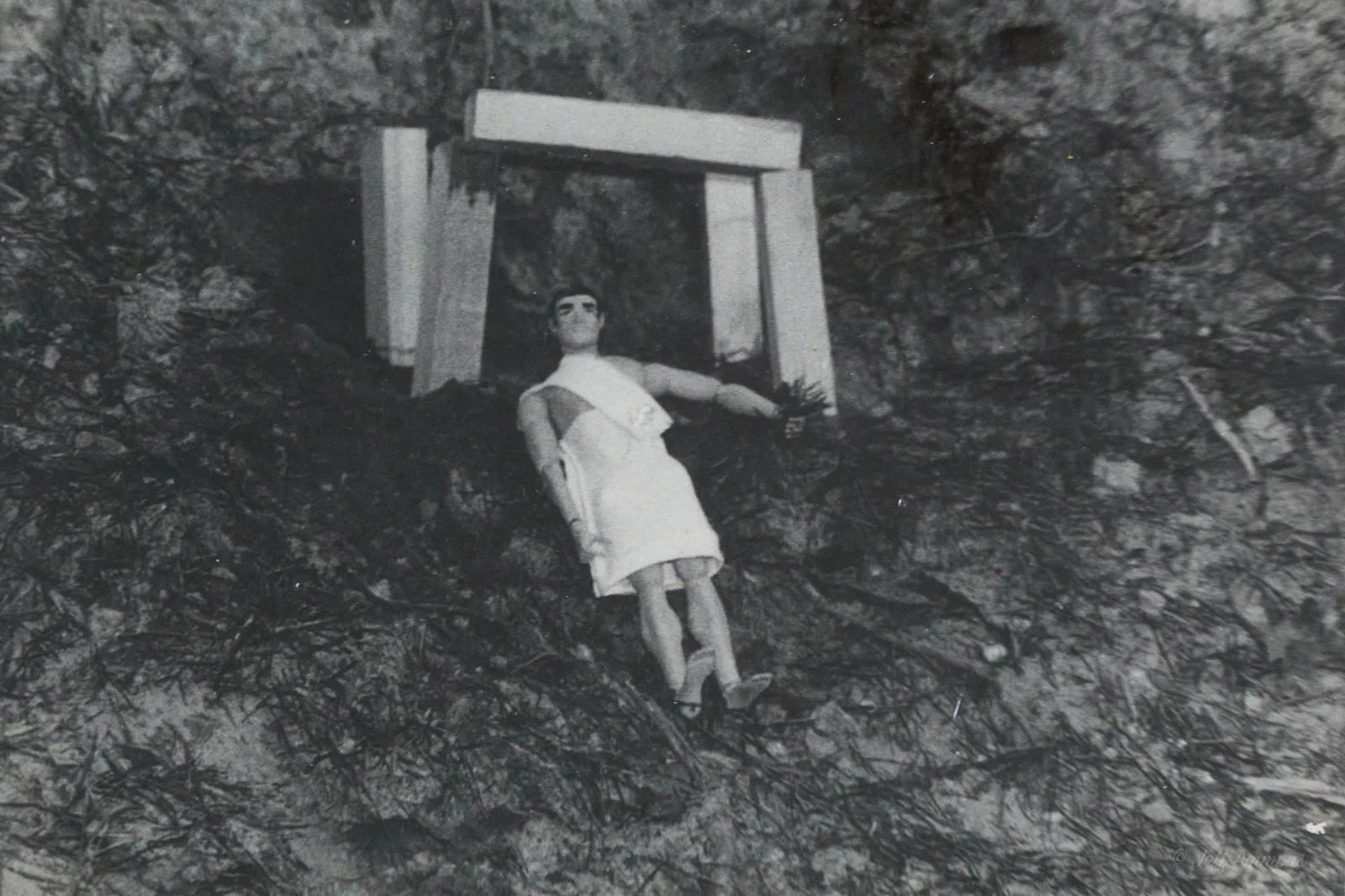 A toga-clad action figure reclines on a rocky hillside in front of a small stone doorway.